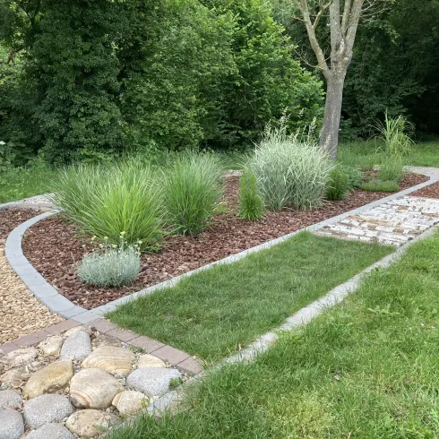 Ein schöner Gartenweg mit verschiedenen Pflanzen und Steinelementen. Der Bereich ist von Grünflächen umgeben und wirkt einladend.