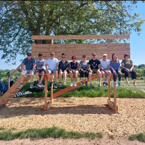 Eine Gruppe von neun Personen sitzt auf einer Holzbank im Freien. Der Himmel ist klar und die Umgebung ist grün und sonnig.