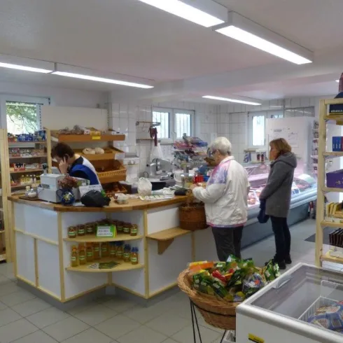 Ein kleines Geschäft mit Regalen, die Lebensmittel und Produkte zeigen. Zwei Kunden stehen an der Kasse, während eine Verkäuferin bedient.