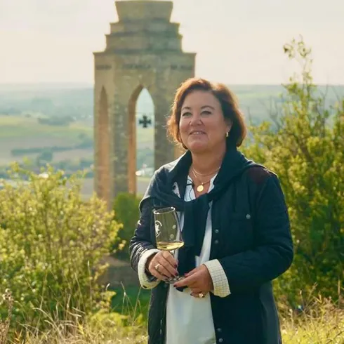 Eine Frau steht lächelnd auf einer Wiese mit einer Aussicht auf eine alte Ruine im Hintergrund. Die Landschaft ist grün und friedlich, was eine angenehme Atmosphäre schafft.
