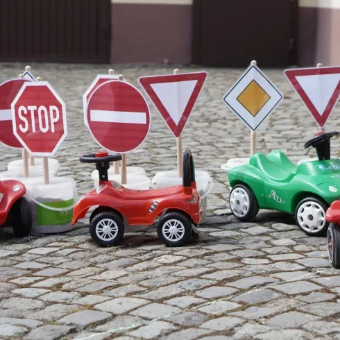 Eine Reihe von Spielzeugautos steht vor verschiedenen Verkehrszeichen auf einem Pflasterboden. Die Autos sind bunt und verkörpern eine kindgerechte Verkehrserziehung.