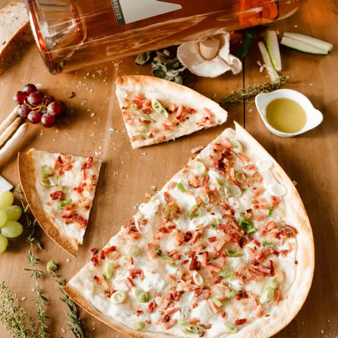 Eine leckere Pizza mit Schinken, Zwiebeln und frischen Kräutern liegt auf einem Holztablett. Daneben stehen ein Glas Wein und verschiedene frische Zutaten.