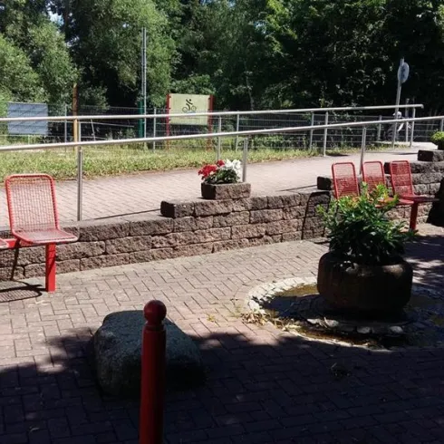 Ein gut gestalteter Außenbereich mit roten Bänken und Pflanzen. Im Hintergrund sind Bäume und eine gepflasterte Fläche sichtbar.