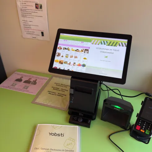Ein moderner Kiosk mit einem Tablet, der eine Willkommensnachricht und verschiedene Produkte anzeigt. Auf dem Tisch liegen Informationsmaterialien und ein Kartenlesegerät.