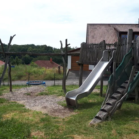 Ein Spielplatz mit einer Rutsche aus Metall und einem Spielhaus aus Holz. Im Hintergrund sind einige Häuser und Bäume zu sehen.