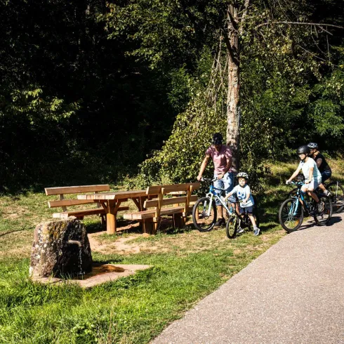 Eine Familie mit Fahrrädern steht an einem Rastplatz. Im Hintergrund sind Bänke und ein Baum zu sehen.