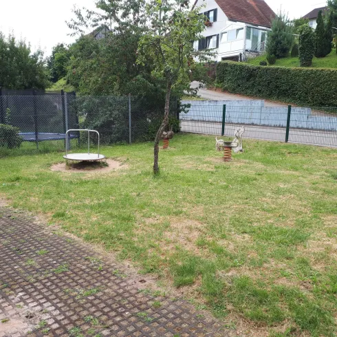 Ein Garten mit einem Spielplatzbereich, der eine Wippe und eine Schaukel umfasst. Die Umgebung ist eingezäunt und von Bäumen und Büschen umgeben.