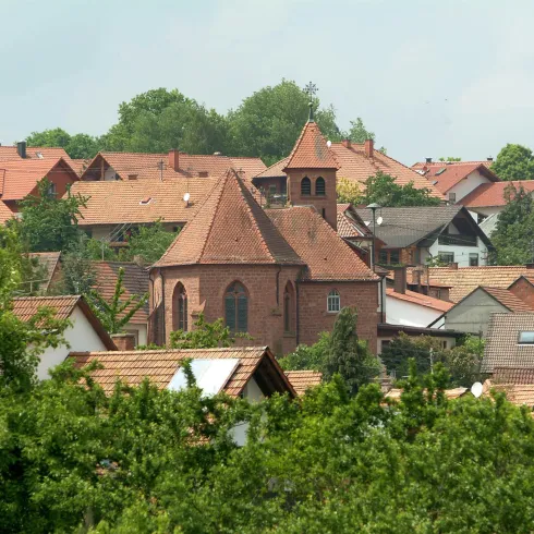 Eine malerische Ansicht eines kleinen Dorfes mit vielen roten Dächern. In der Mitte steht eine auffällige Kirche umgeben von Bäumen.