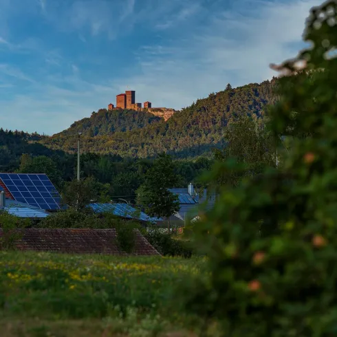 Eine alte Burg thront auf einem Hügel hinter einem ländlichen Landschaftsbild. Im Vordergrund sind grüne Felder und Solarpanels zu sehen.