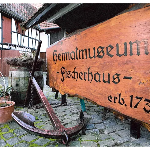 Ein rustikales Schild mit der Aufschrift „Heimatmuseum Fischernhaus“ und einem Anker. Im Hintergrund sind historische Gebäude und Pflanzen zu sehen.