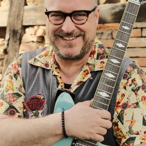 Ein lächelnder Mann mit Brille hält eine blaue Gitarre. Er trägt ein farbenfrohes Hemd und steht vor einer rustikalen Hintergrundkulisse.