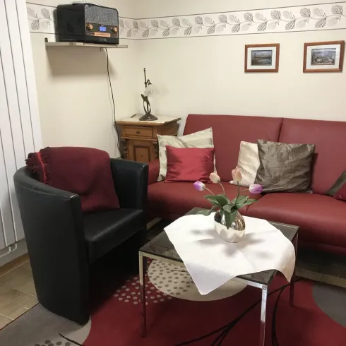 Ein gemütliches Wohnzimmer mit einem roten Sofa und einem schwarzen Sessel. Auf dem Tisch liegt eine weiße Decke mit einer Blumenvase.