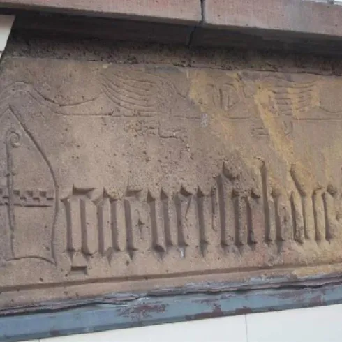 Eine Steinplatte mit eingravierten Buchstaben und einem dekorativen Muster. Die Schrift ist schwer leserlich und zeigt historische Handwerkskunst.