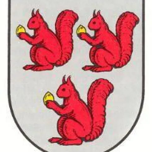 Drei rote Eichhörnchen, die jeweils eine gelbe Nuss halten, auf einem grauen Wappen. Das Wappen hat eine runde Form und einfache Farben.