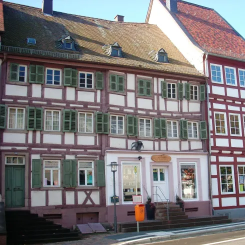 Ein historisches Fachwerkhaus mit grünen Fensterläden und einer schönen Fassade. Nebenan steht ein weiteres traditionelles Gebäude mit roten und weißen Elementen.