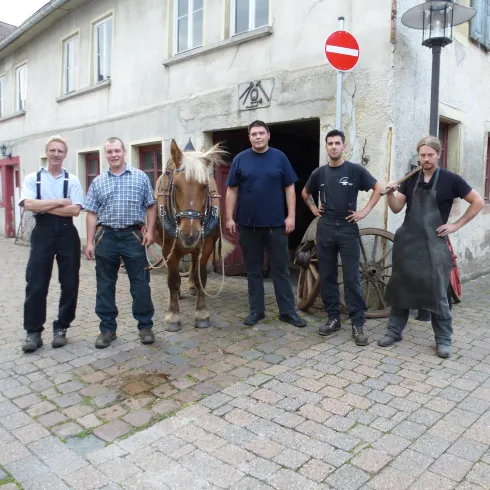 Fünf Personen stehen auf einem gepflasterten Platz neben einem Pferd. Im Hintergrund ist ein altes Gebäude und ein Verkehrsschild zu sehen.