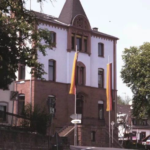 Ein historisches Gebäude mit einem spitzen Dach und Fahnen. Die Straße führt sanft bergab in eine ruhige Umgebung.