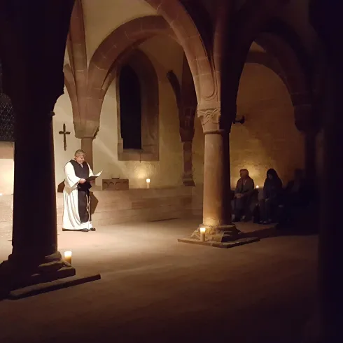 Ein dunkler Raum mit hohen, bogenförmigen Säulen. In der Mitte steht eine Person mit einem Buch, während andere Personen im Hintergrund aufmerksam lauschen.