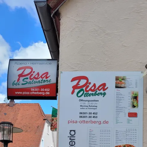 Ein Restaurant-Schild mit der Aufschrift "Pisa" und einer Speisekarte für Pizzas und andere Speisen. Das Schild ist an einer Wand angebracht und hat eine ansprechende Gestaltung.