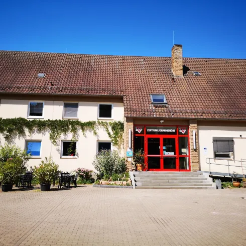 Ein modernes Gebäude mit einer roten Eingangstür und grünen Pflanzen. Der Himmel ist klar und blau.