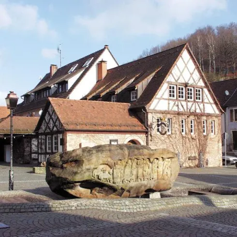 Ein traditionelles Fachwerkhaus mit roten Dächern und einem großen, kunstvoll gestalteten Stein im Vordergrund. Die Umgebung ist ruhig und einladend, umgeben von Bäumen.