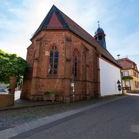 Eine alte Kirche aus roten Ziegeln steht an einer Straße. Im Hintergrund sind weitere Gebäude und Bäume sichtbar.