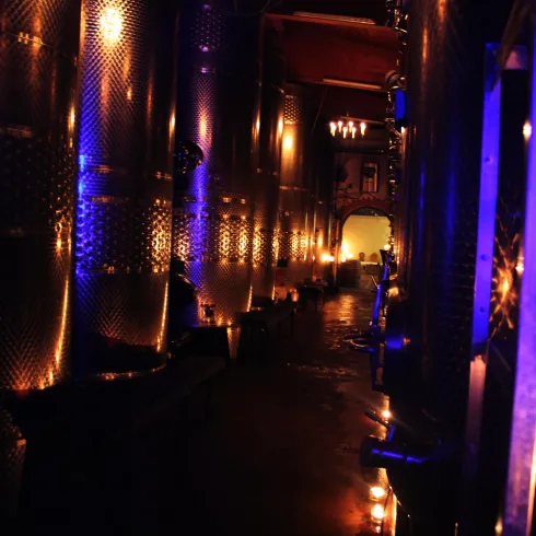 Ein dunkler Gang in einem Weinkeller, beleuchtet von Kerzen und sanften blauen Lichtern. Die Wände sind mit großen, glänzenden Metallbehältern ausgestattet.