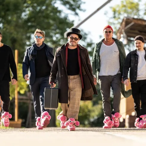 Eine Gruppe von fünf Männern läuft selbstbewusst auf einem Weg. Sie tragen ungewöhnliche pinkfarbene Hausschuhe und unterschiedliche stylische Outfits.