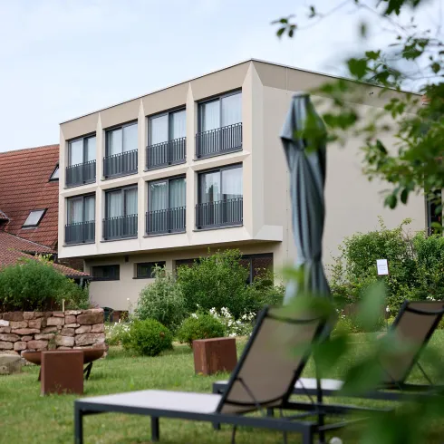 Ein modernes Hotelgebäude mit mehreren Fenstern und Balkonen. Im Vordergrund sind Liegen und eine grüne Wiese zu sehen.