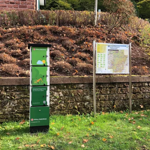 Ein Informationsstand mit grüner Farbgebung steht neben einer Landschaftsfläche. Im Hintergrund ist ein Informationsschild mit einer Karte und Daten zu sehen.