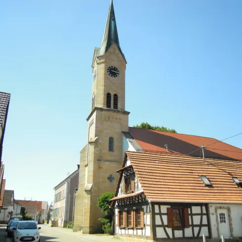 Eine alte Kirche mit einem hohen Turm steht in einer ruhigen Straße. Umgeben von traditionellen Gebäuden und klarem blauen Himmel.