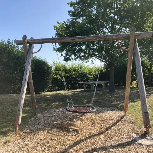 Eine Schaukel aus Holz steht auf einem Spielplatz, umgeben von Gras und Bäumen. Der Himmel ist klar und es ist ein sonniger Tag.