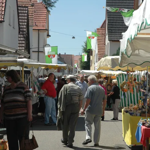 Ein belebter Markt mit Ständen und Menschen, die umherlaufen. Die Stände bieten verschiedene Waren unter Sonnenschirmen an.