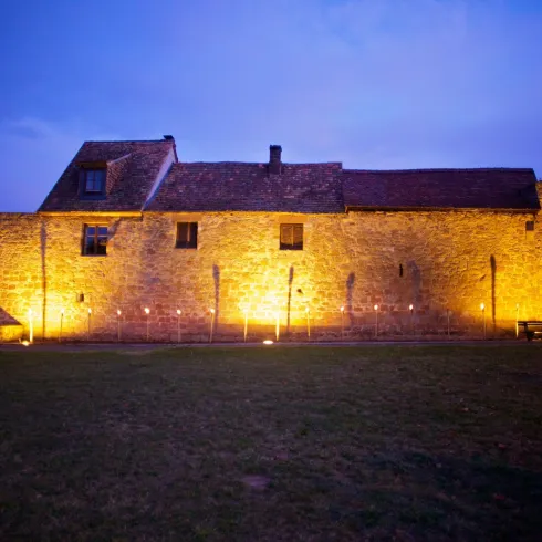 Ein altes Gebäude aus Stein, beleuchtet bei Dämmerung. Die Umgebung ist grün und offen.