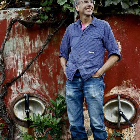 Ein lächelnder Mann in Gummistiefeln steht vor einer alten Wand, die mit Pflanzen bewachsen ist. Hinter ihm sind große, runde Metallbehälter zu sehen.