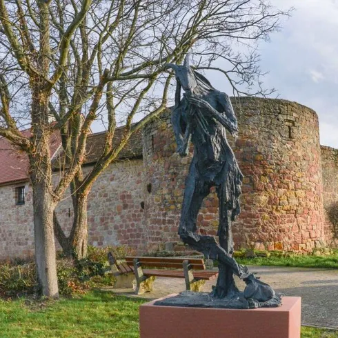 Eine Statue eines kranken Mannes steht in einem Park. Im Hintergrund ist eine alte Mauer und ein Baum zu sehen.