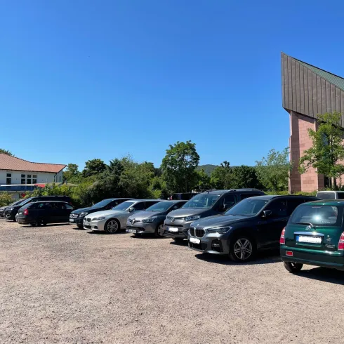 Ein Parkplatz mit mehreren geparkten Autos und einem klaren blauen Himmel. Im Hintergrund ist ein modernes Gebäude und einige Bäume zu sehen.