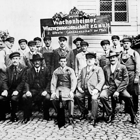 Eine Gruppe von Männern in historischen Arbeitskleidern steht vor einem Schild mit der Aufschrift „Wachonheimer Wasserzubehörgesellschaft“. Die Aufnahme scheint aus einer vergangenen Zeit zu stammen und zeigt eine Verbindung zur lokalen Industrie.