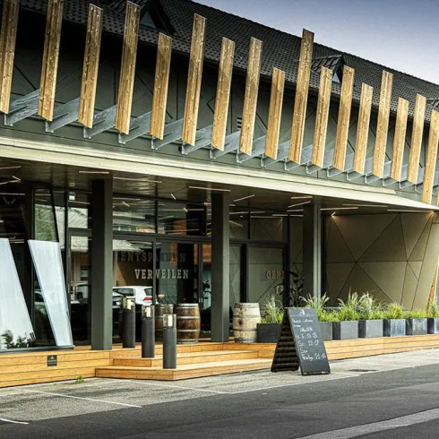 Ein modernes Gebäude mit Holz- und Glaselementen. Vor dem Eingang stehen Pflanzen und Fässer, die eine einladende Atmosphäre schaffen.