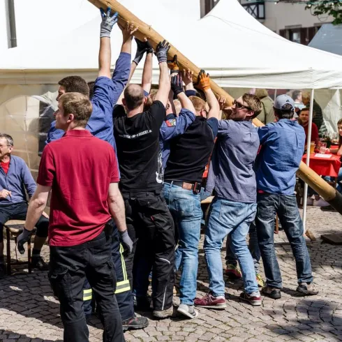 Eine Gruppe von Männern hebt gemeinsam einen langen Balken an. Im Hintergrund sind Tische und andere Personen bei einer festlichen Veranstaltung zu sehen.