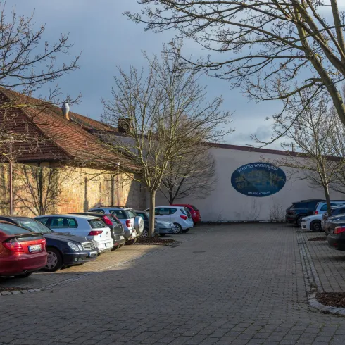 Ein Parkplatz mit mehreren Autos und kahlen Bäumen. Im Hintergrund ist ein Gebäude mit einem Wandbild zu sehen.