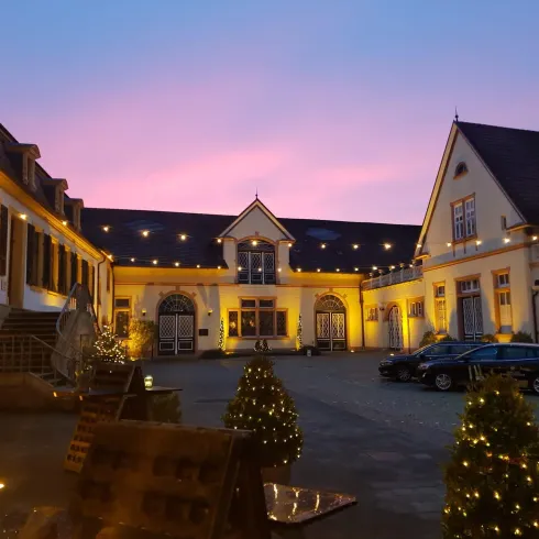 Ein wunderschöner Innenhof mit festlicher Beleuchtung und einem beeindruckenden Gebäude im Hintergrund. Der Himmel strahlt in sanften Lila- und Rosatönen.