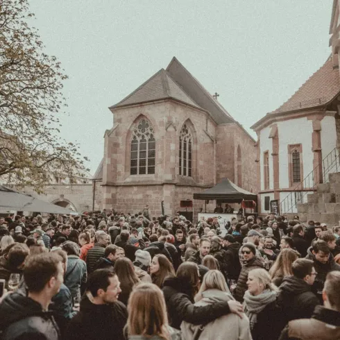 Eine große Menschenmenge versammelt sich vor einer historischen Kirche. Es herrscht eine festliche Atmosphäre mit Bäumen und Ständen im Hintergrund.