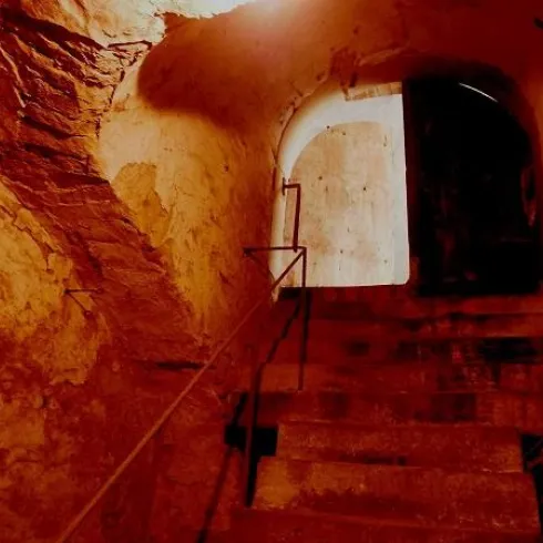 Eine alte steinerne Treppe führt zu einer dunklen Tür. Das Licht fällt schwach auf die Wände und vermittelt eine geheimnisvolle Stimmung.