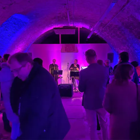 Ein lebhaftes Event in einem künstlerisch beleuchteten Raum mit einer Band auf der Bühne. Menschen unterhalten sich und genießen die Atmosphäre.