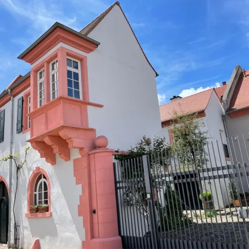 Ein charmantes Haus mit einer pinken Fensterlaibung und grünen Fensterläden. Im Hintergrund sind weitere Gebäude und ein blauer Himmel zu sehen.