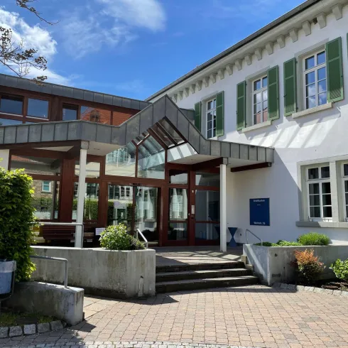 Ein modernes Gebäude mit einer großen Glasfassade und einem einladenden Eingangsbereich. Die Umgebung ist grün und gut gepflegt.