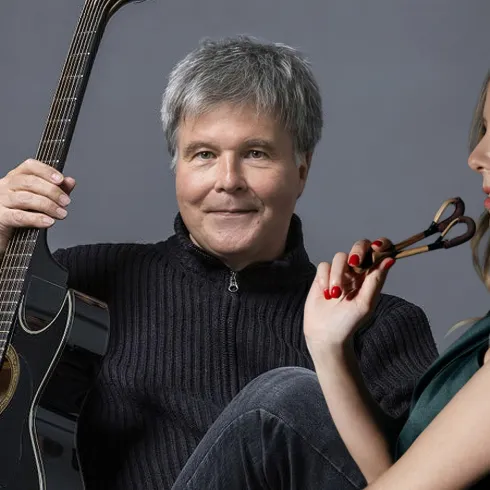 Ein Mann mit einer Gitarre sitzt auf einem Sessel und lächelt. Neben ihm sitzt eine Frau, die nachdenklich einen Pinsel hält.
