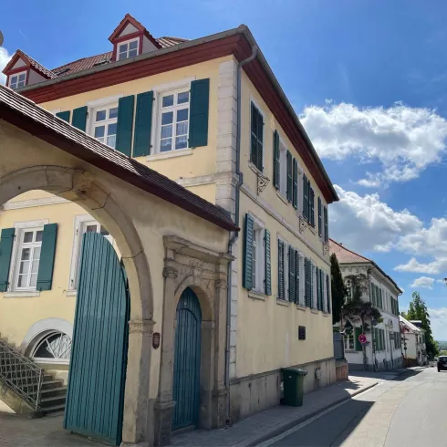 Ein klassisches Gebäude mit grünen Fensterläden und einem großen Bogen. Die Straße ist ruhig und es gibt einige Bäume und Wolken im Hintergrund.
