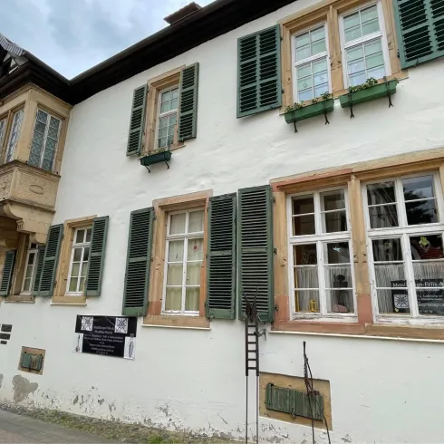 Ein historisches Gebäude mit weißen Wänden und grünen Fensterläden. Es hat eine markante Architektur mit einem Erker an der Vorderseite.